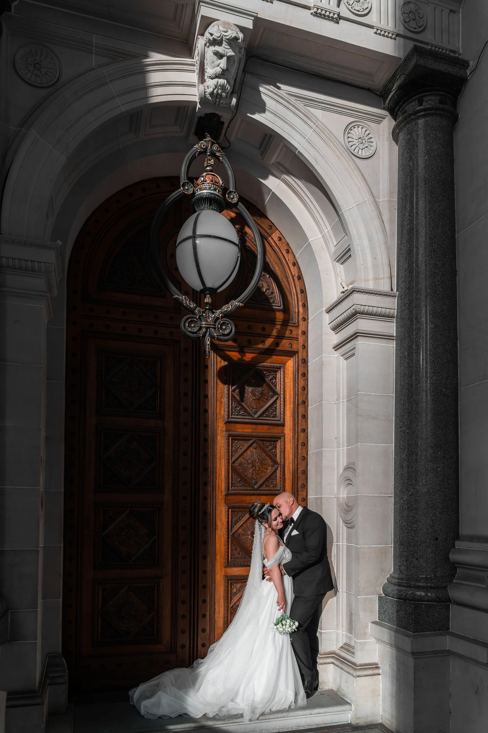 Bride and groom sharing a romantic kiss under an ornate stone archway with a grand wooden door.