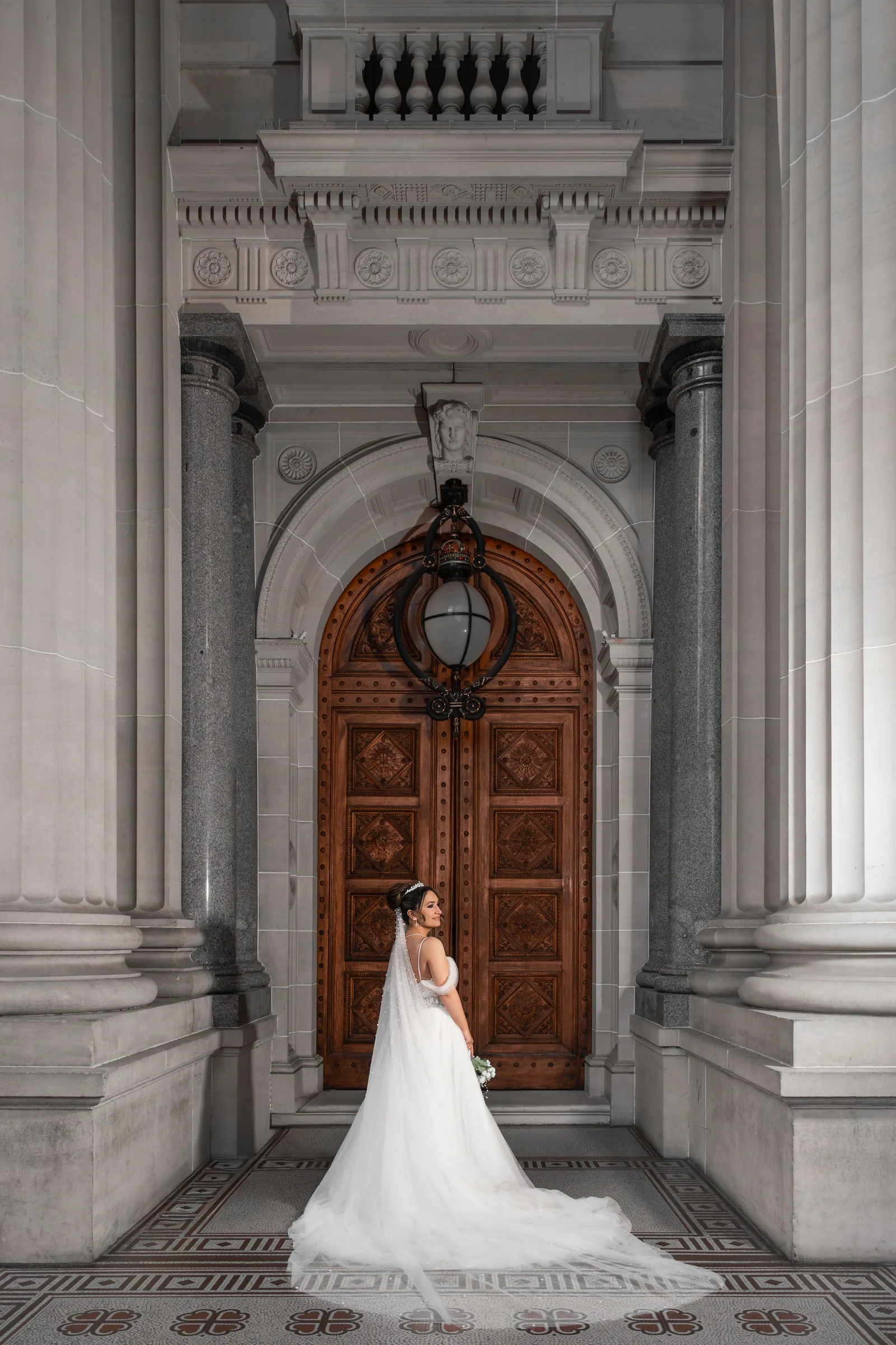 Bride in white gown and veil poses before ornate wooden arched door flanked by stone columns.