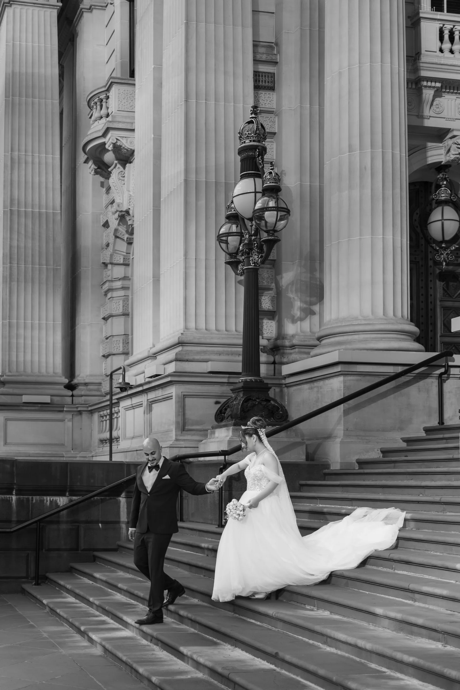 Black and white photo of bride and groom walking down grand stone steps with tall columns and vintage lamp post.