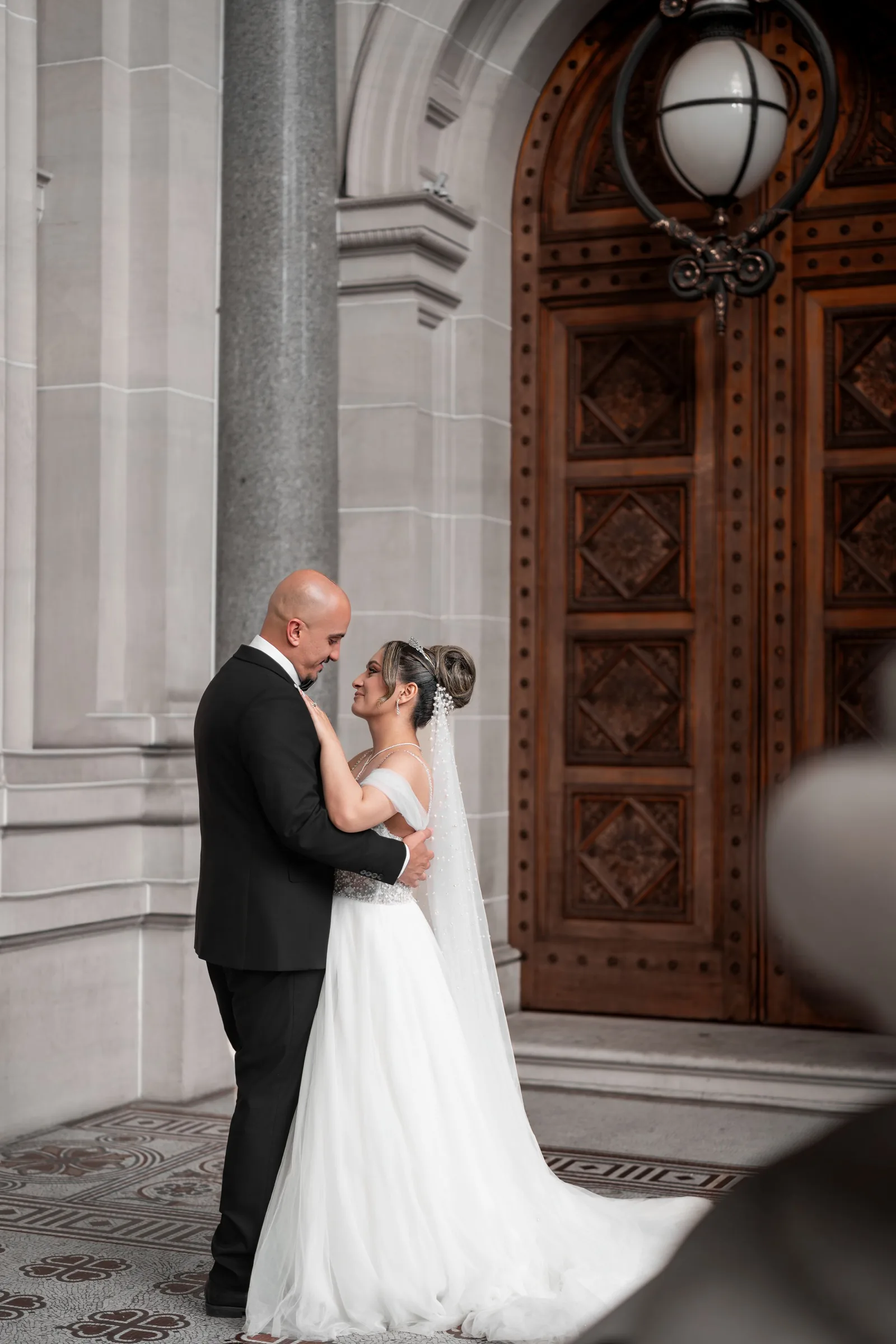 Bride and groom share a romantic embrace outside ornate wooden doors of an elegant stone building on their wedding day.