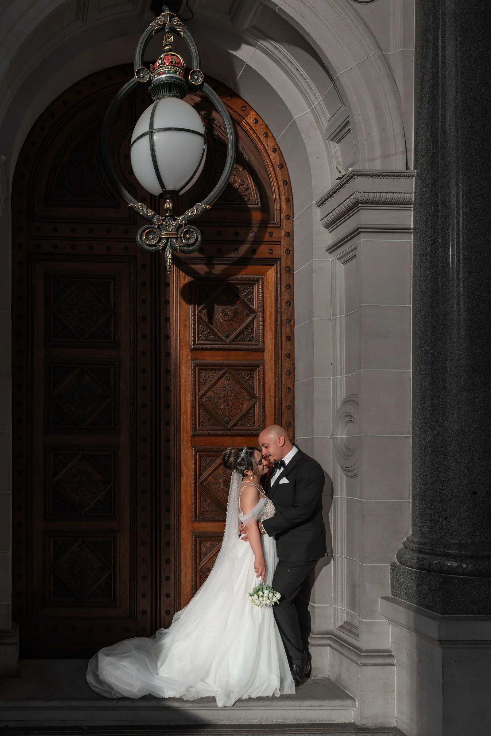Bride and groom share a romantic kiss under an ornate lantern at a grand stone building entrance.