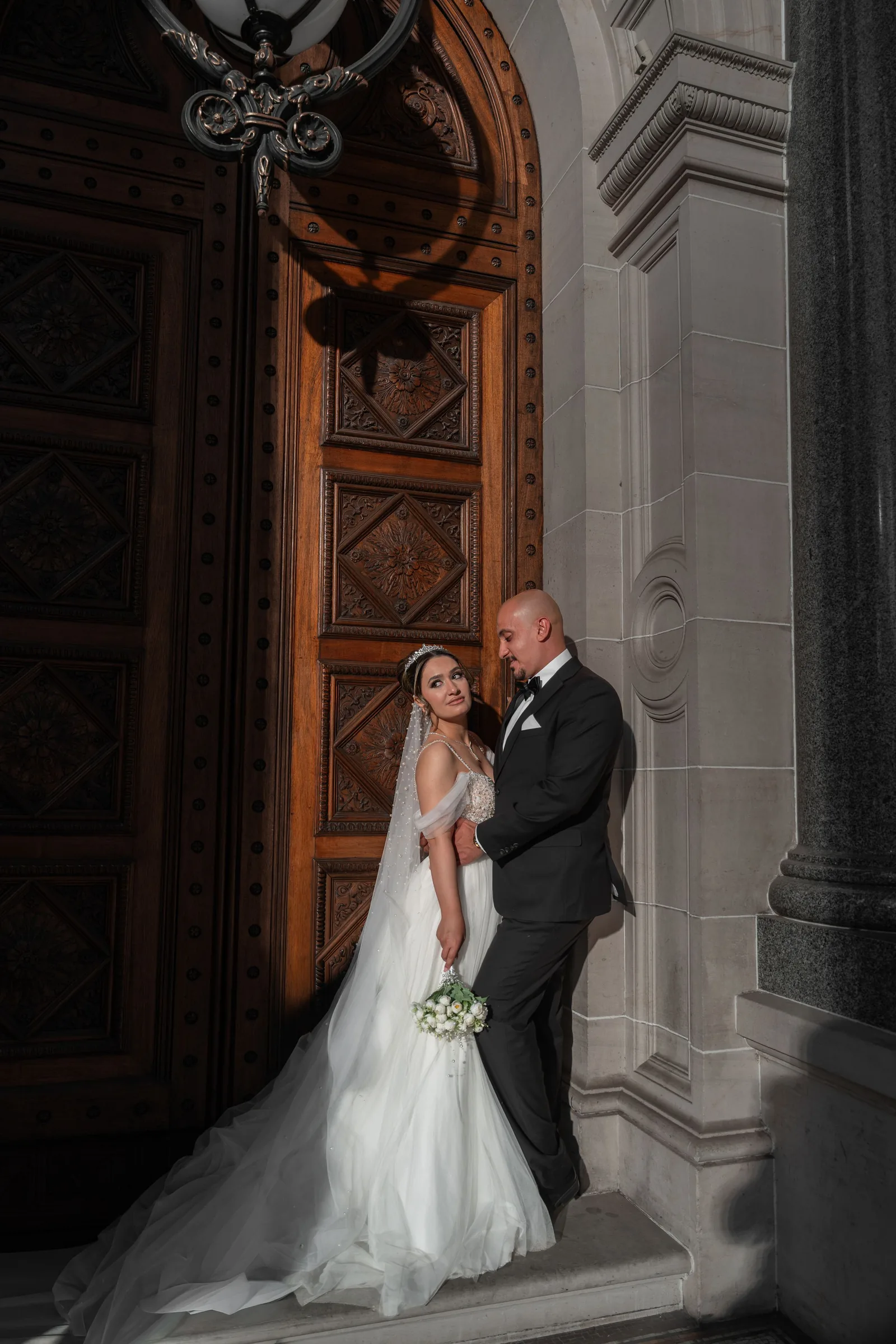 Bride in flowing white gown and veiled headpiece embraces groom beside ornate wooden cathedral doors.