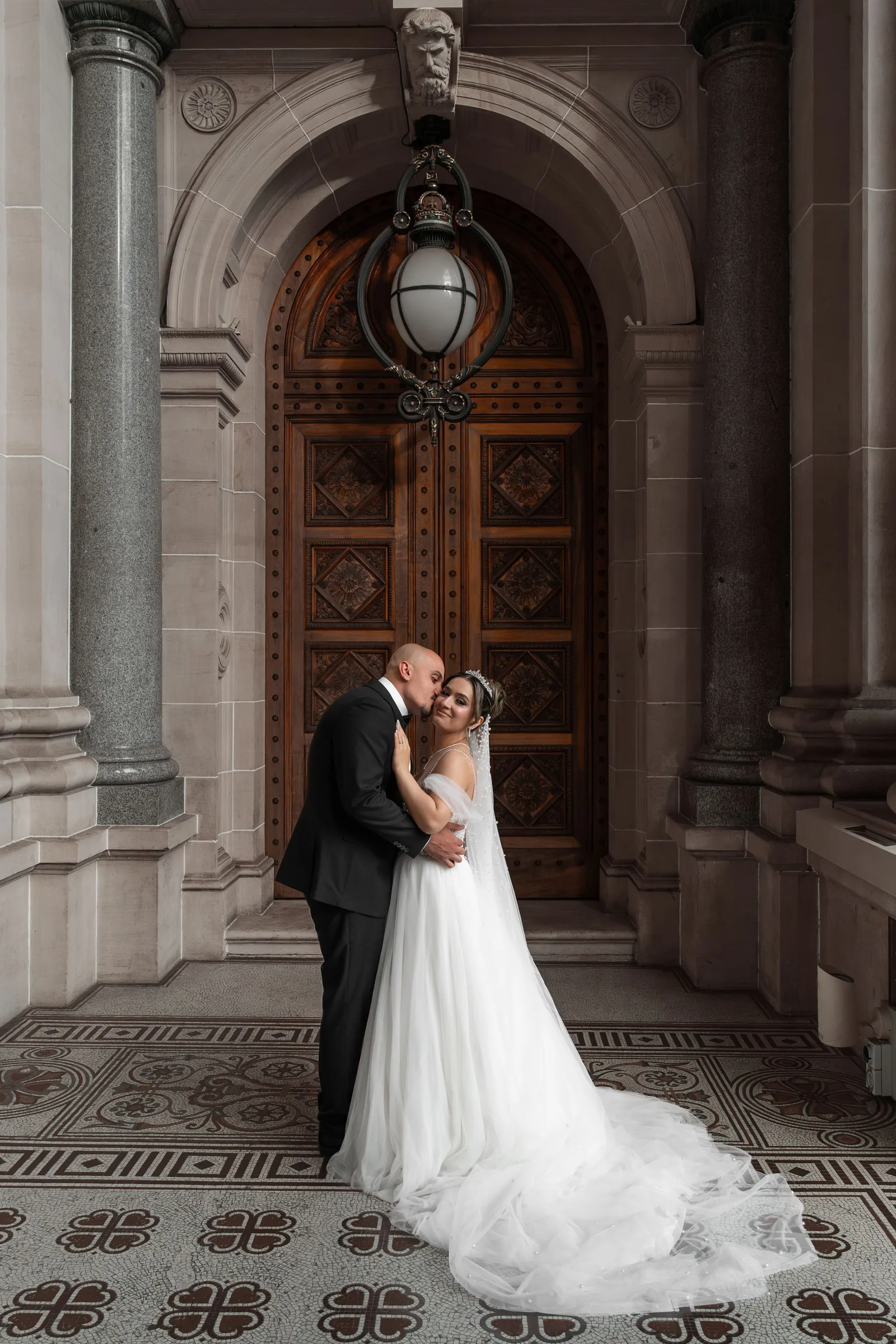 Wedding couple embracing tenderly in front of an elegant carved wooden door with decorative tile floor.