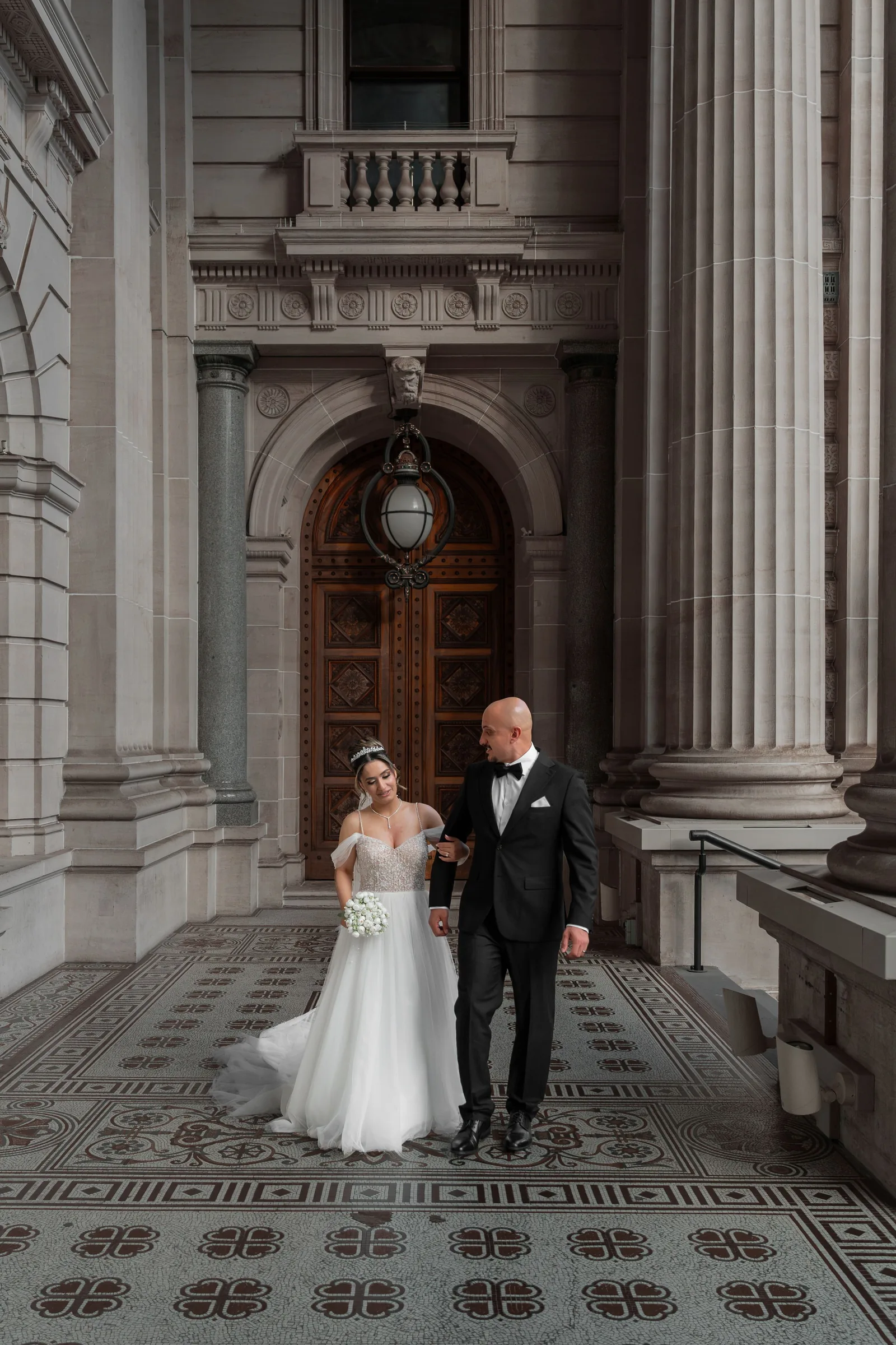 Elegant bride in floral gown and groom in tuxedo walk hand in hand before ornate neoclassical doors.