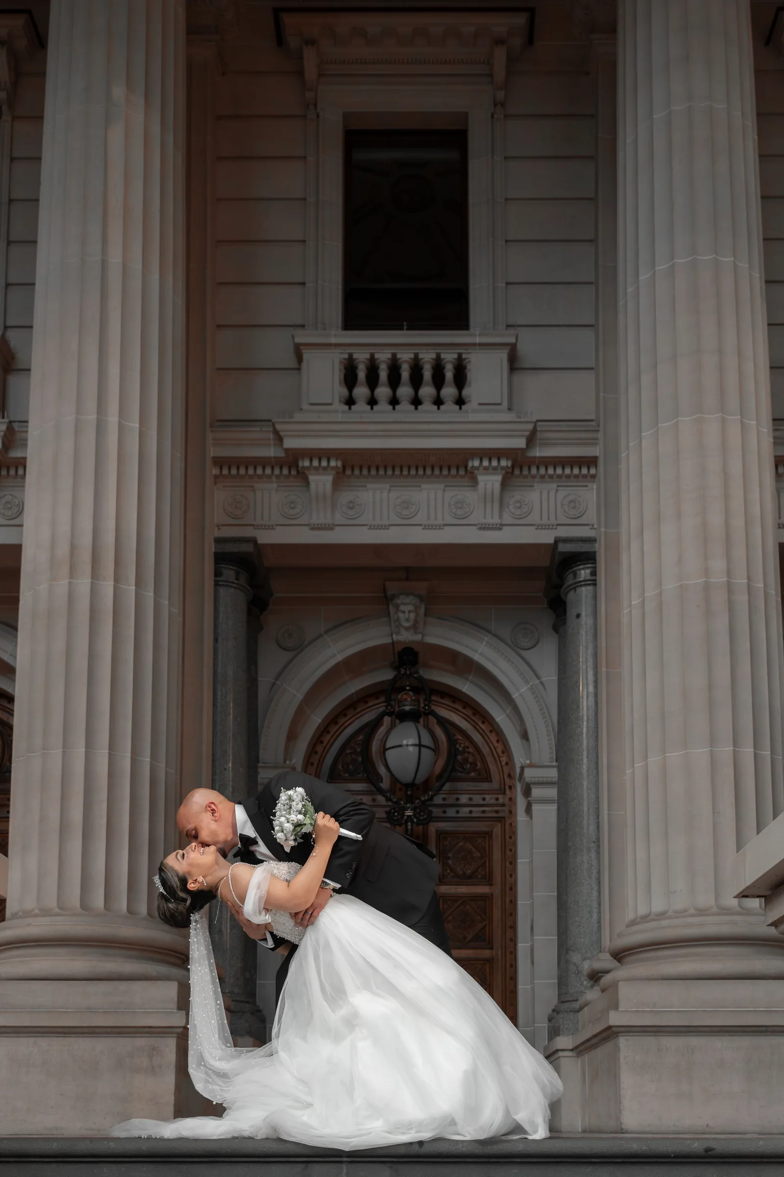 Groom dips bride dramatically before grand neoclassical building with tall stone columns and arched doorway.