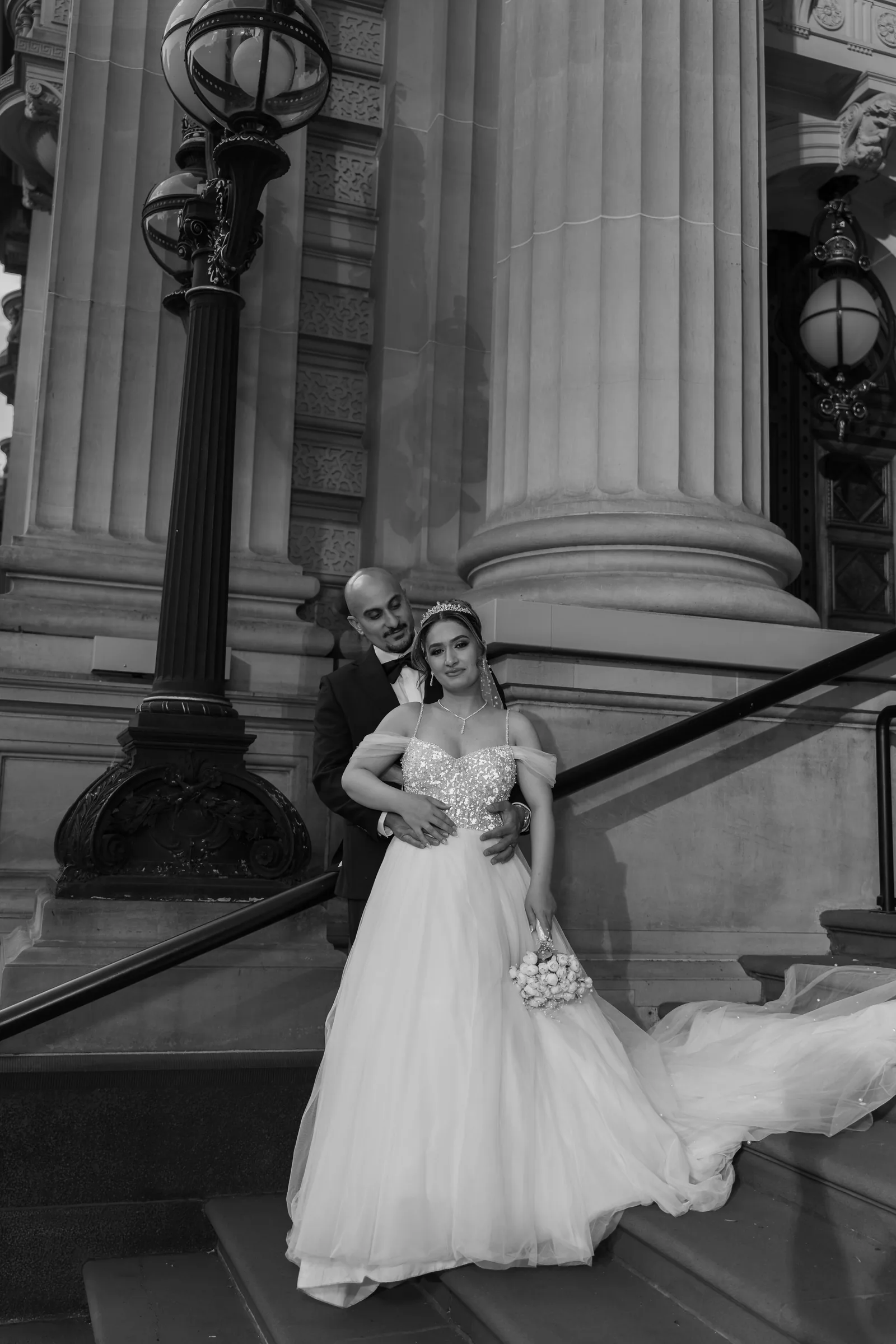 Black and white photo of bride and groom posing on steps beside tall classical columns.