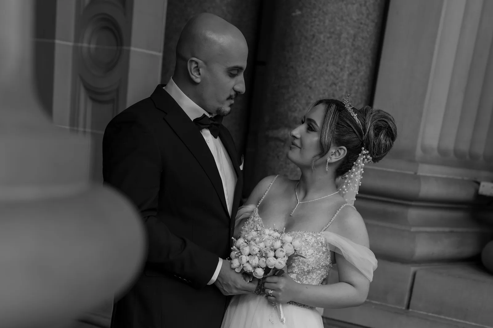 Black and white wedding photo of bride holding bouquet and groom in tuxedo sharing intimate moment by stone columns.