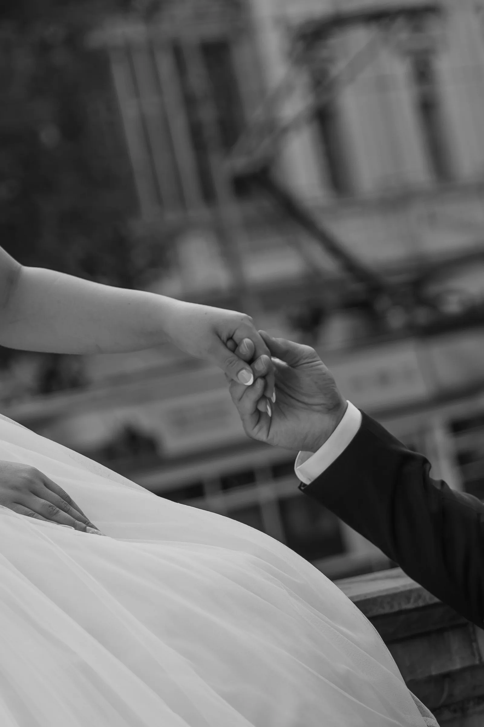 Black and white photo of a bride and groom gently holding hands, with a blurred building in the background.