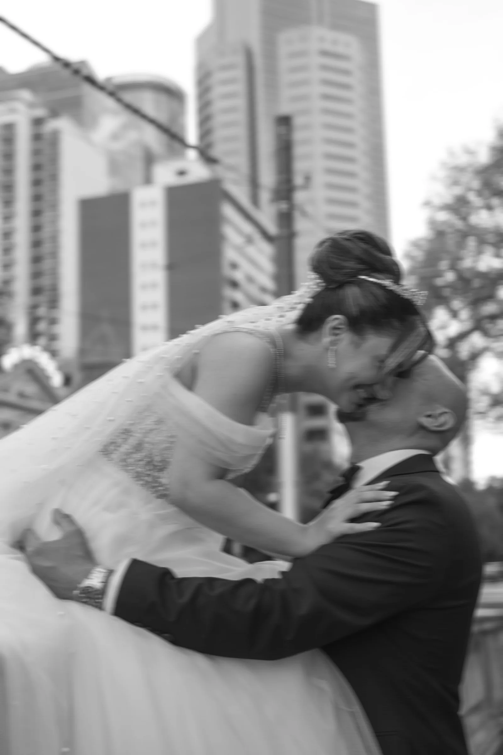 Bride and groom share a passionate kiss in black and white photo with city skyscrapers in background.