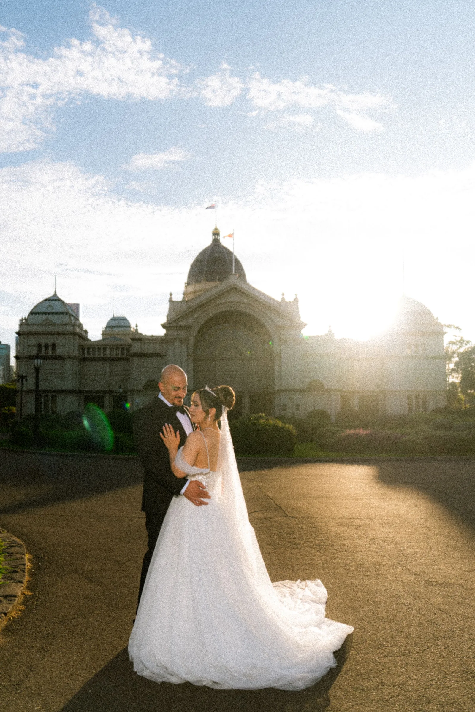 Bride and groom share a romantic embrace in front of a grand historic building at golden hour.