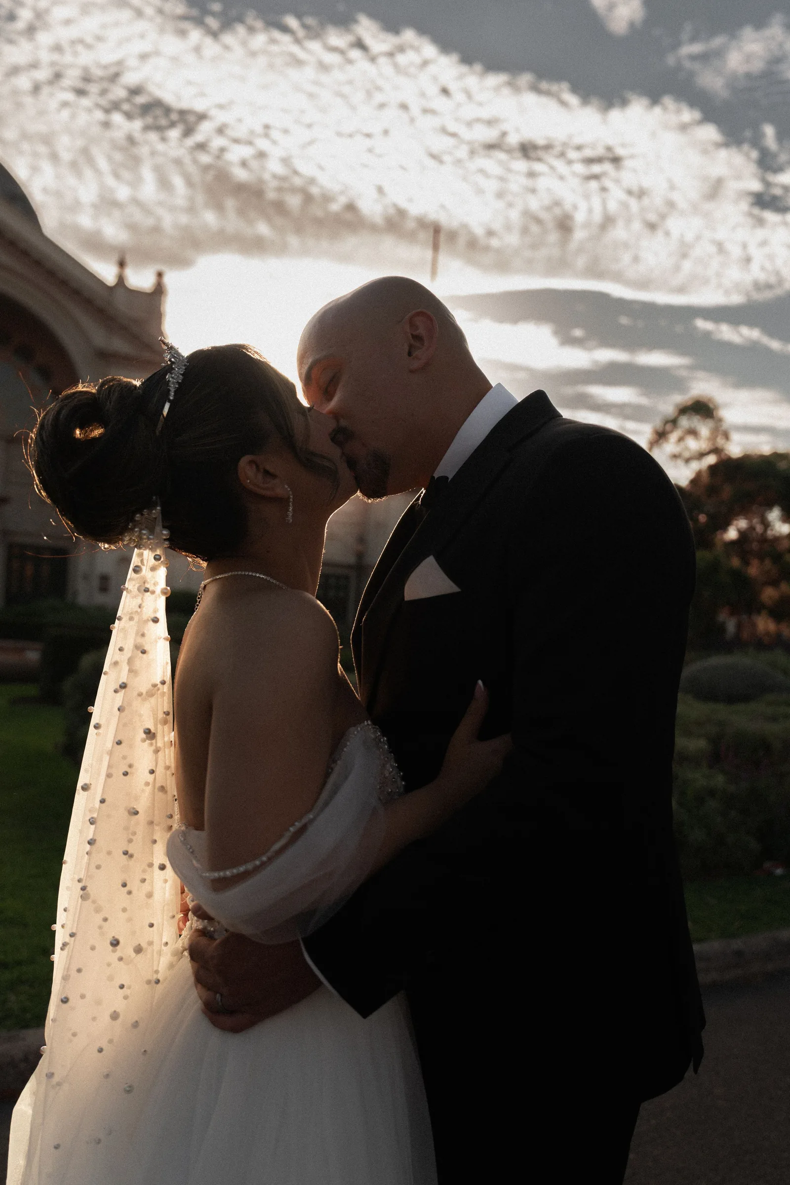 Bride and groom sharing a romantic kiss at sunset, bride in white gown with veil, groom in black tuxedo.
