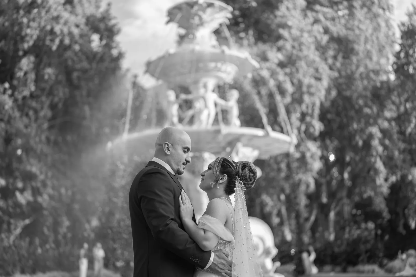 Black and white wedding photo of couple embracing near ornate fountain in garden.