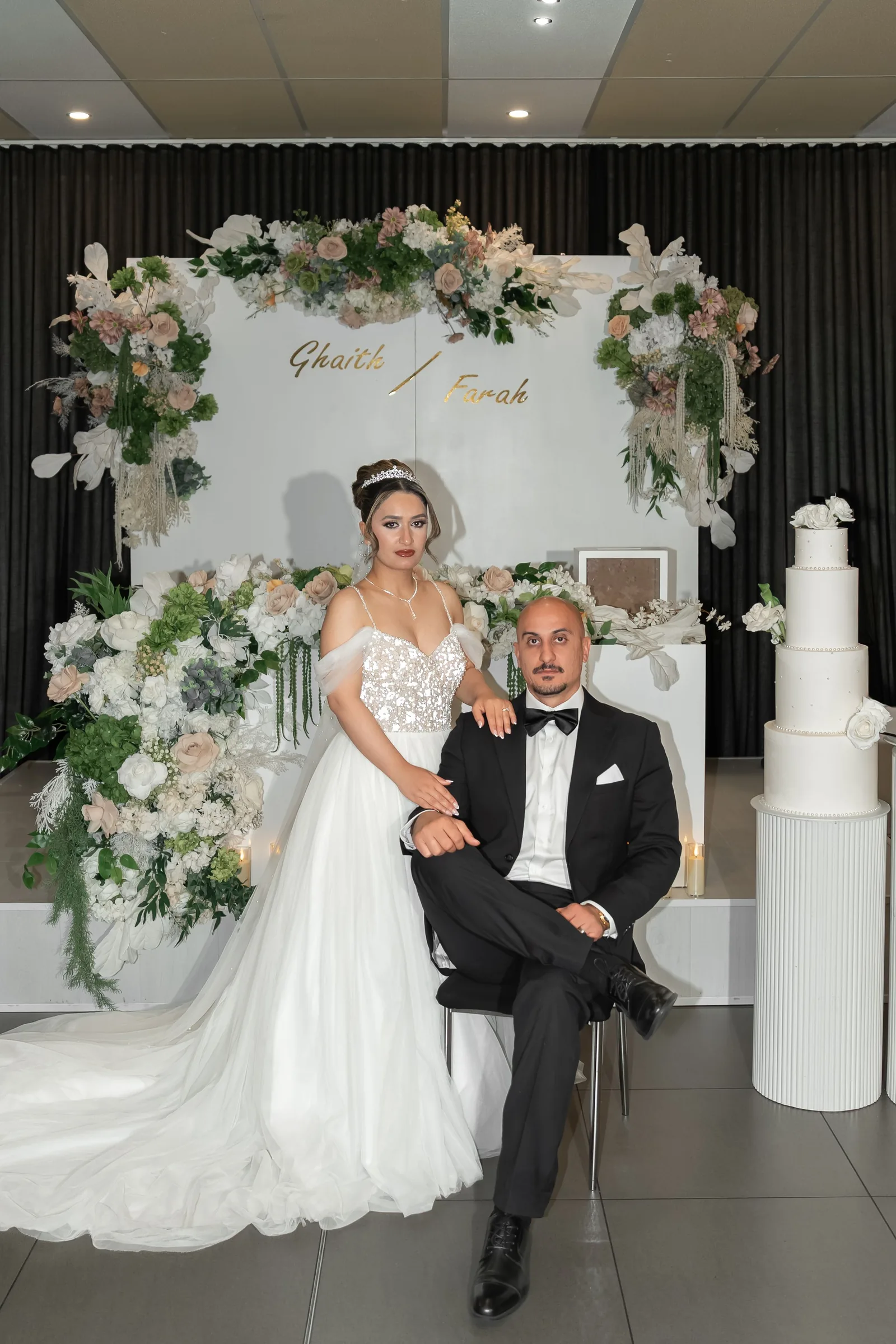Bride in white gown and groom in black tuxedo pose before floral arch backdrop at elegant wedding reception.