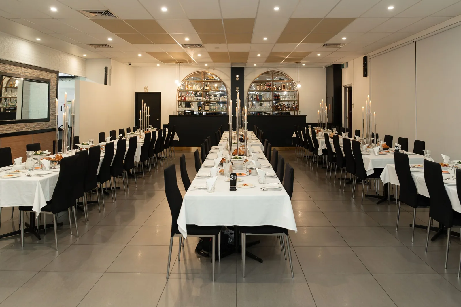Elegant wedding reception venue with long white-clothed tables, black chairs, and candlelit centerpieces in a spacious dining room.
