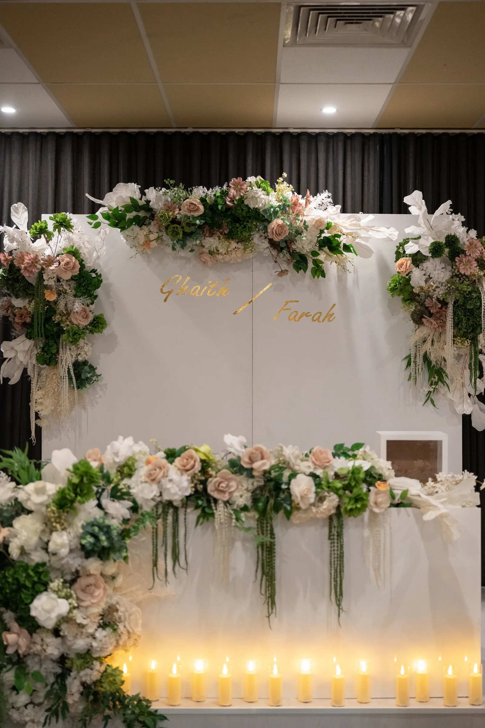 Elegant wedding backdrop with blush roses, white florals, greenery, gold names, and candles on a sweetheart table.
