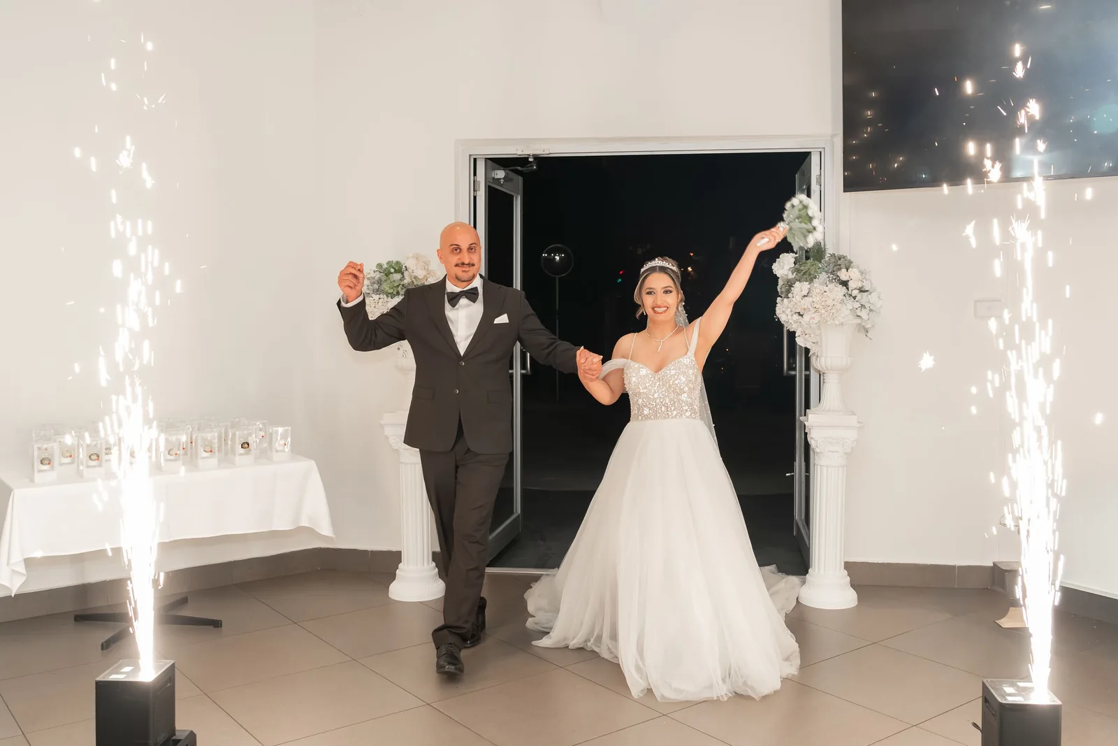 Bride and groom make grand entrance with cold spark fountains at elegant white wedding reception venue.
