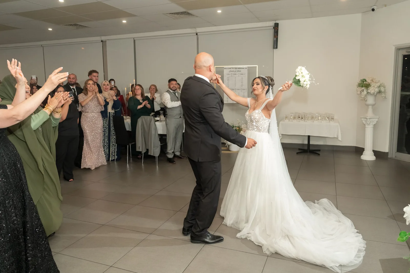 Bride in white gown and groom dancing joyfully at wedding reception while guests clap and celebrate.