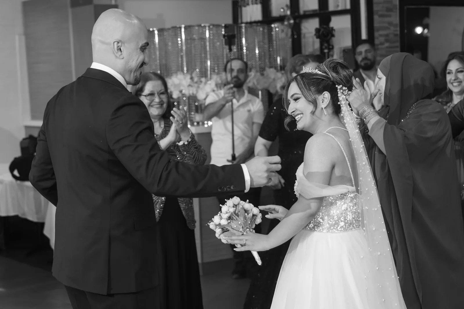 Smiling bride holding bouquet reaches for grooms hand as wedding guests celebrate in black and white photo.