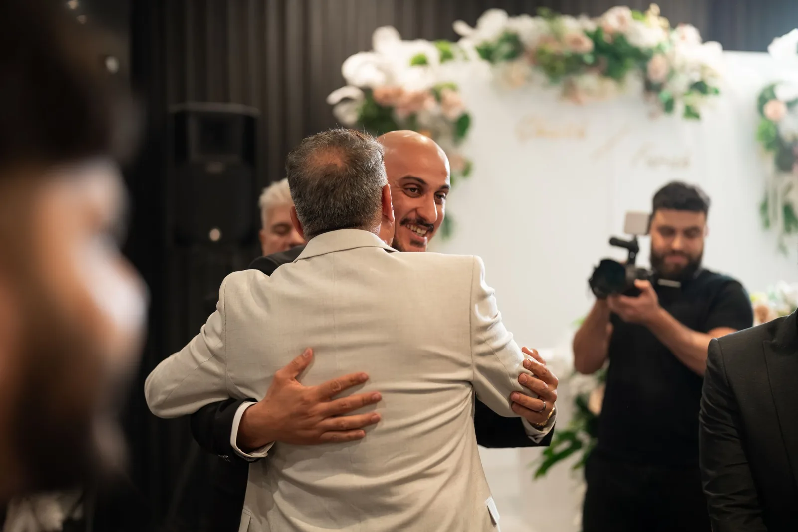 Groom in white suit sharing a warm embrace with a guest at a wedding reception with floral arch backdrop.