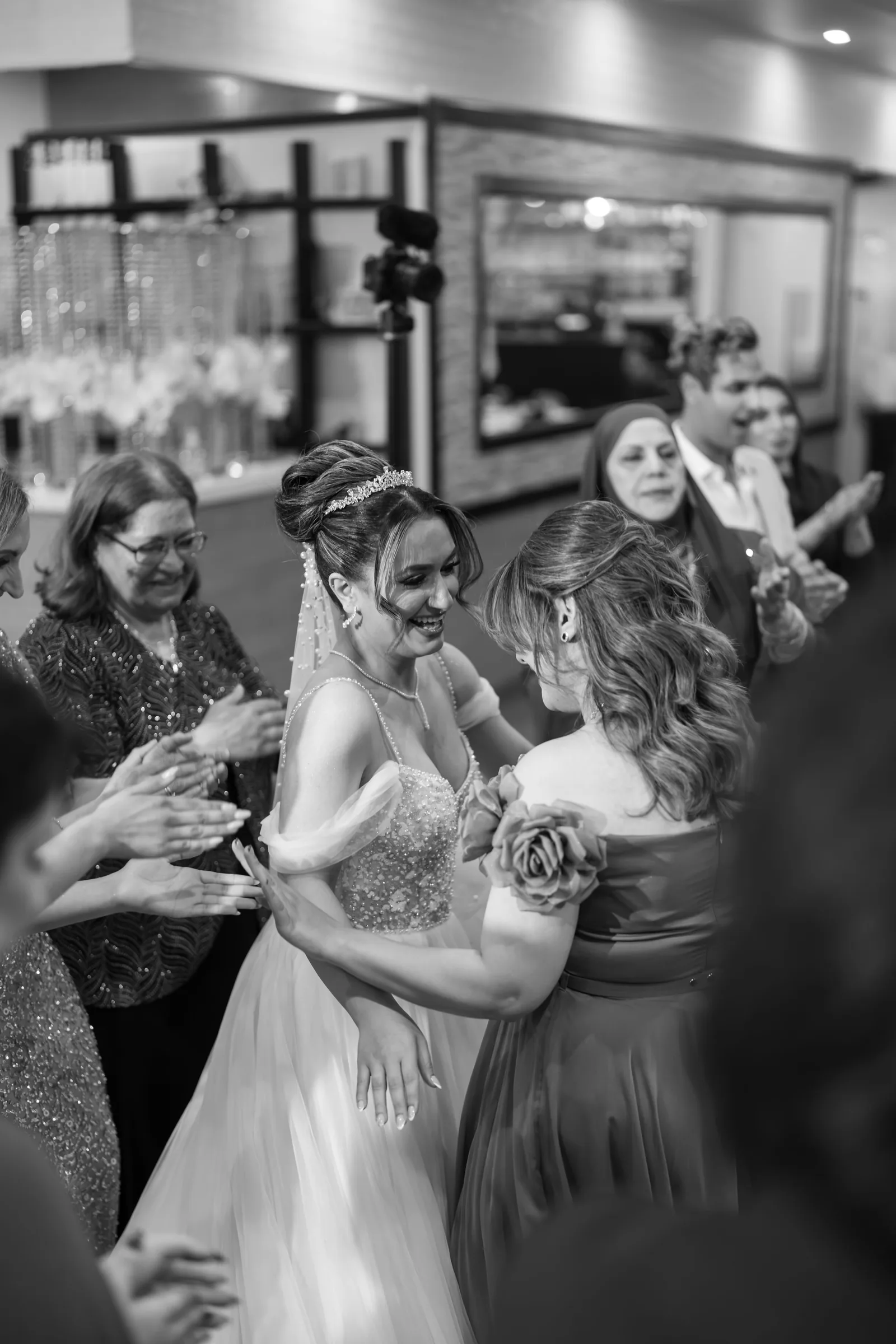 Bride in beaded gown and veil hugs bridesmaid while wedding guests celebrate and clap at reception in black and white photo.