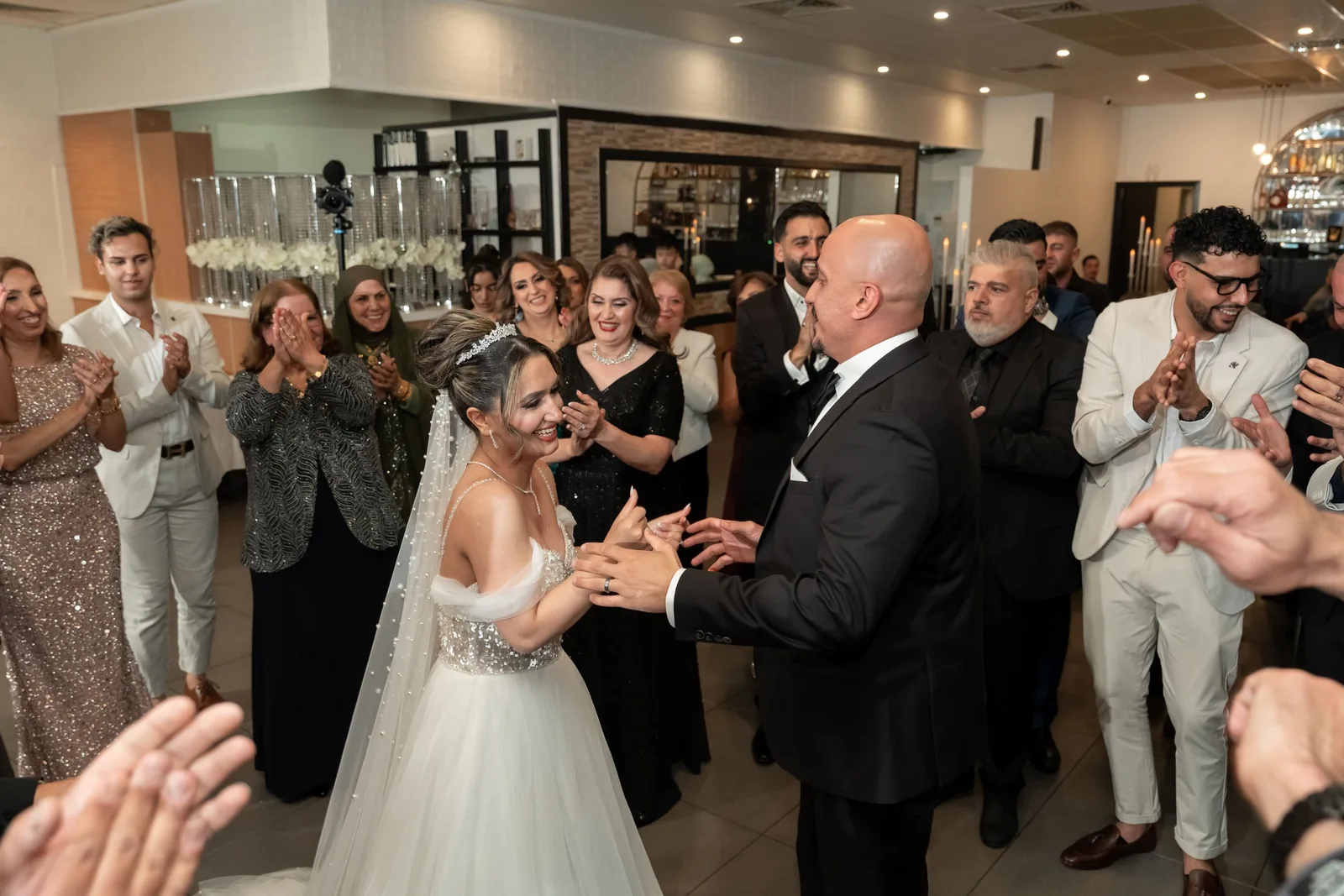 Bride and groom share first dance surrounded by clapping wedding guests in elegant reception venue.