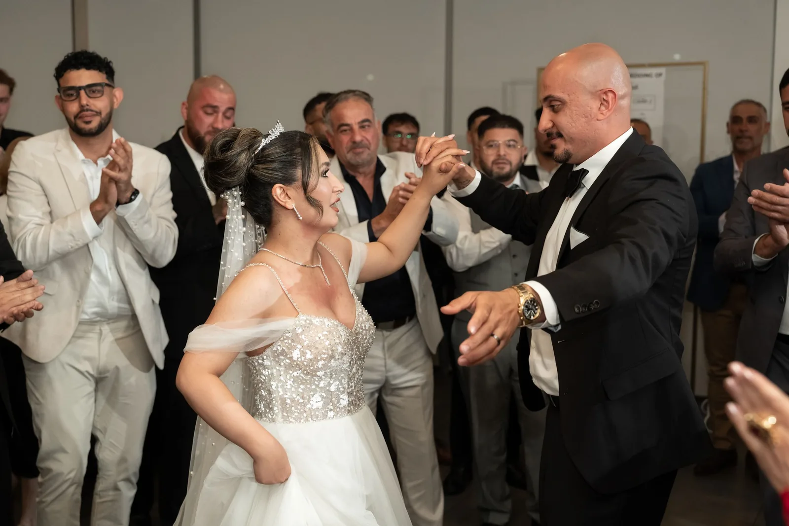 Bride and groom dance together as smiling guests applaud at lively wedding reception celebration.