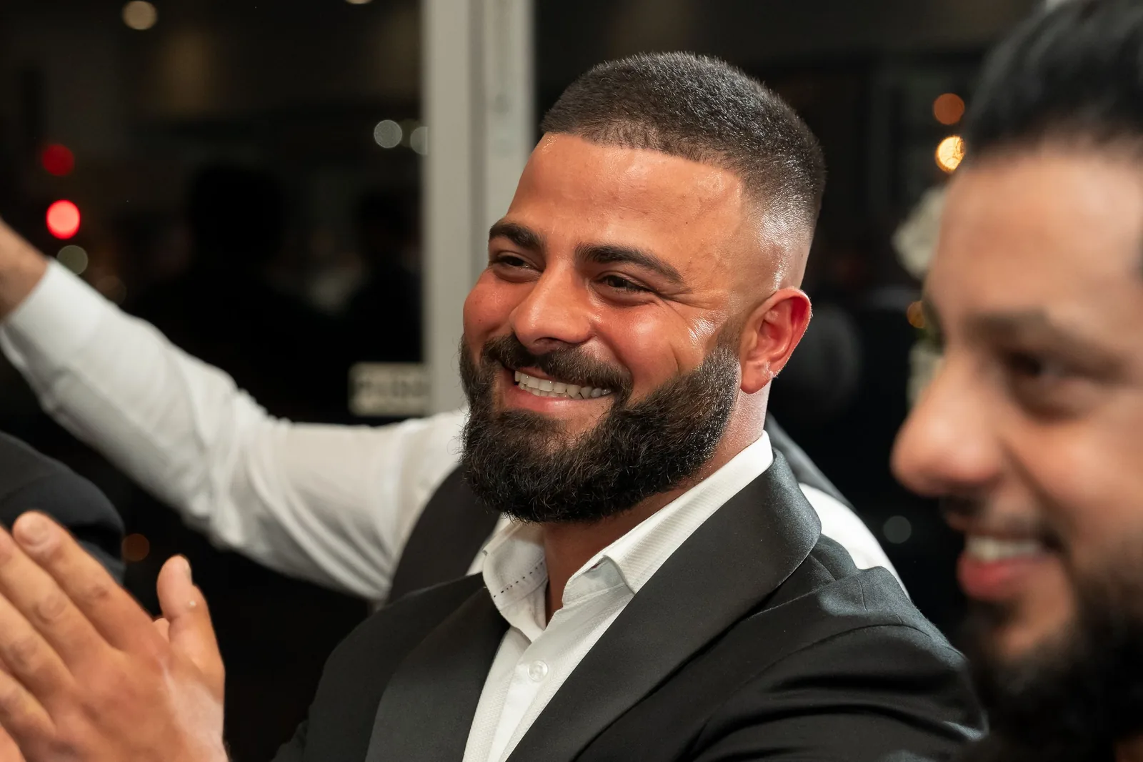 Smiling bearded man in a black suit at a social event, surrounded by guests in a festive atmosphere.