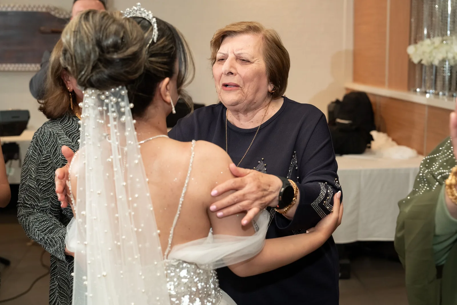 Elderly woman warmly embracing a bride in a beaded wedding gown and veil at a wedding reception.