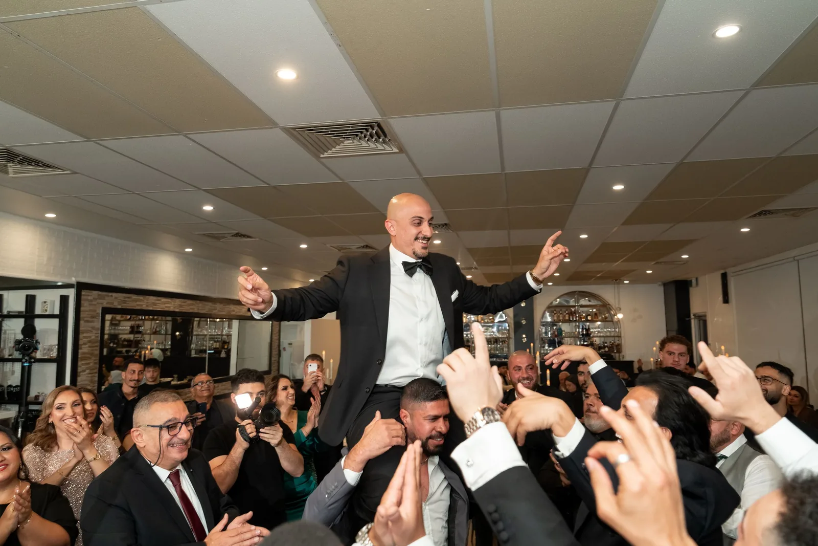 Groom in tuxedo lifted by cheering wedding guests during lively reception celebration.