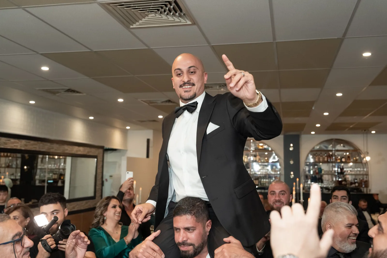 Groom in tuxedo celebrates atop guests shoulders pointing up during lively wedding reception party.