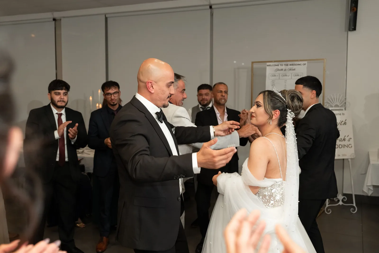 Bride in white gown dances with bald man in tuxedo at wedding reception while guests clap and celebrate.