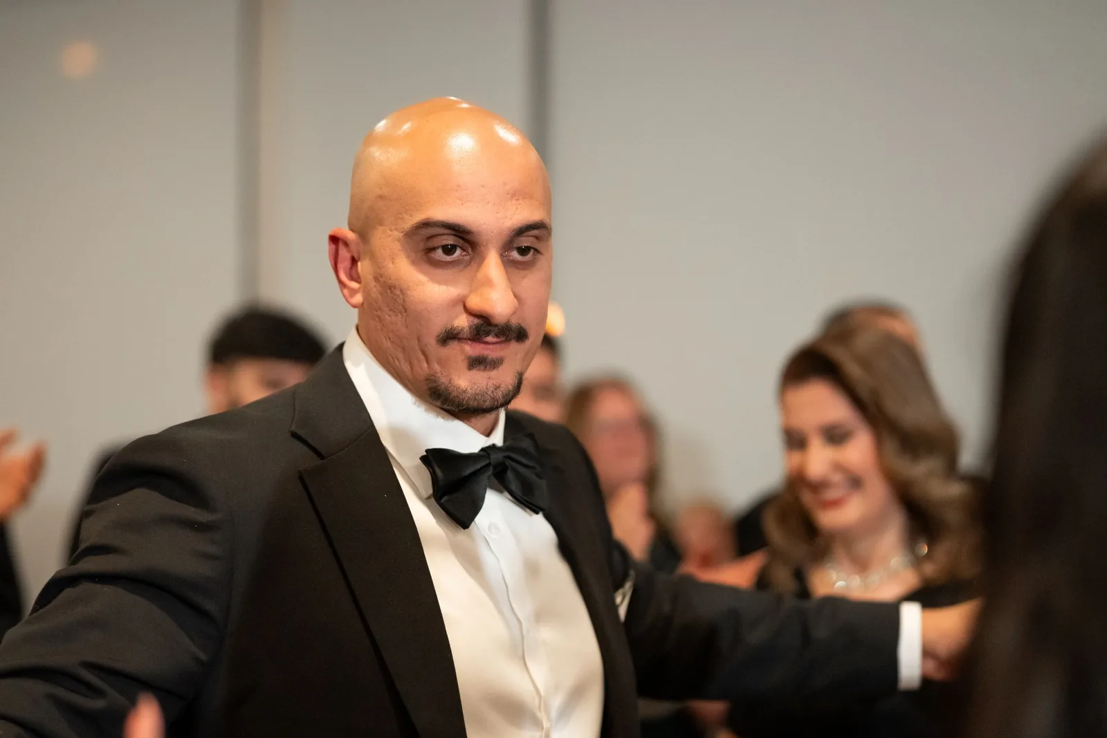 Elegant man in black tuxedo with bow tie smiles at formal wedding reception event.