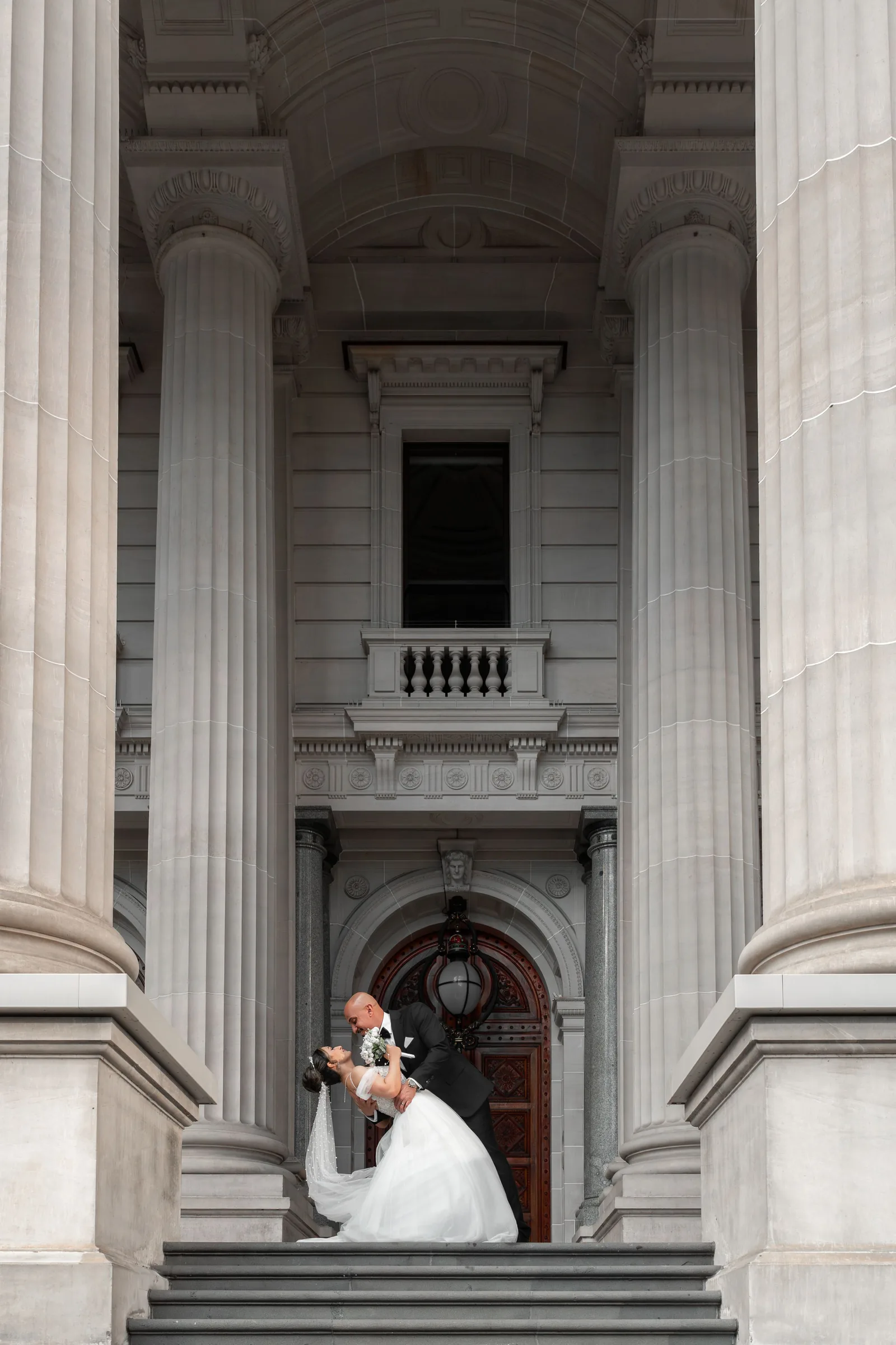 Bride and groom share a romantic dip kiss on grand neoclassical building steps with tall stone columns.