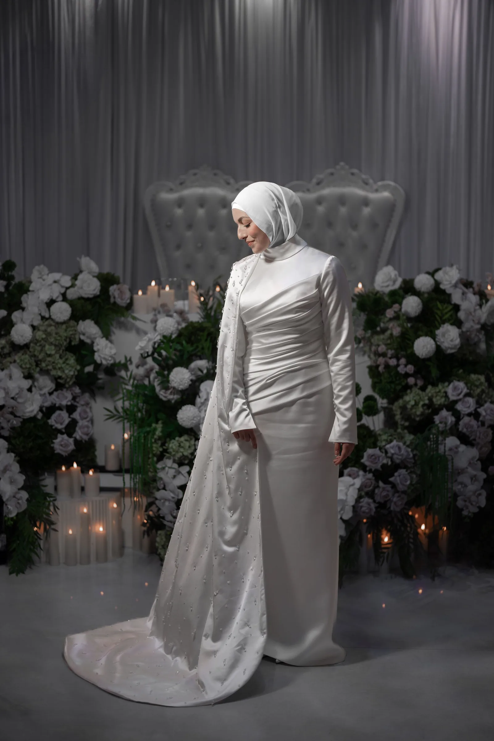 Elegant wedding couple poses in white attire against floral backdrop with ambient lighting.