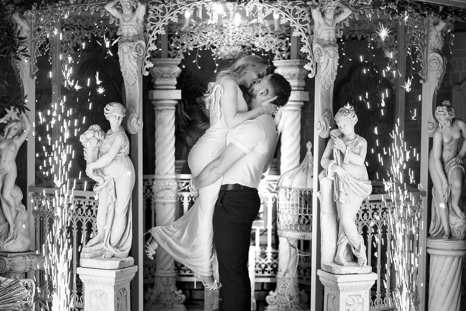 Black and white photo of the couple embracing between classical statues and columns as sparklers fall behind them