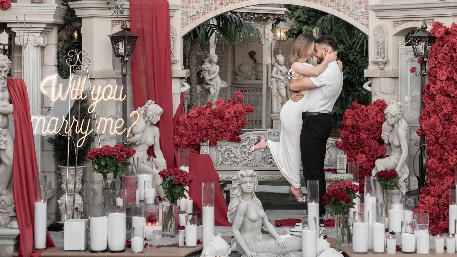 Proposal setup with a Will you marry me neon sign, red roses and candles as the couple kiss on the steps