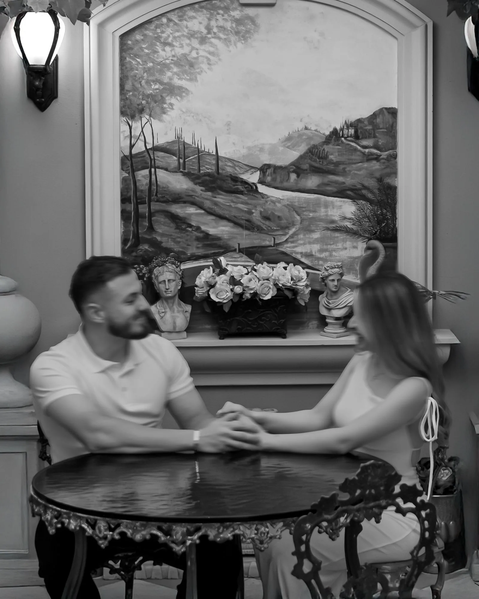 Black and white photo of the couple holding hands across a small round table beneath a painted landscape mural