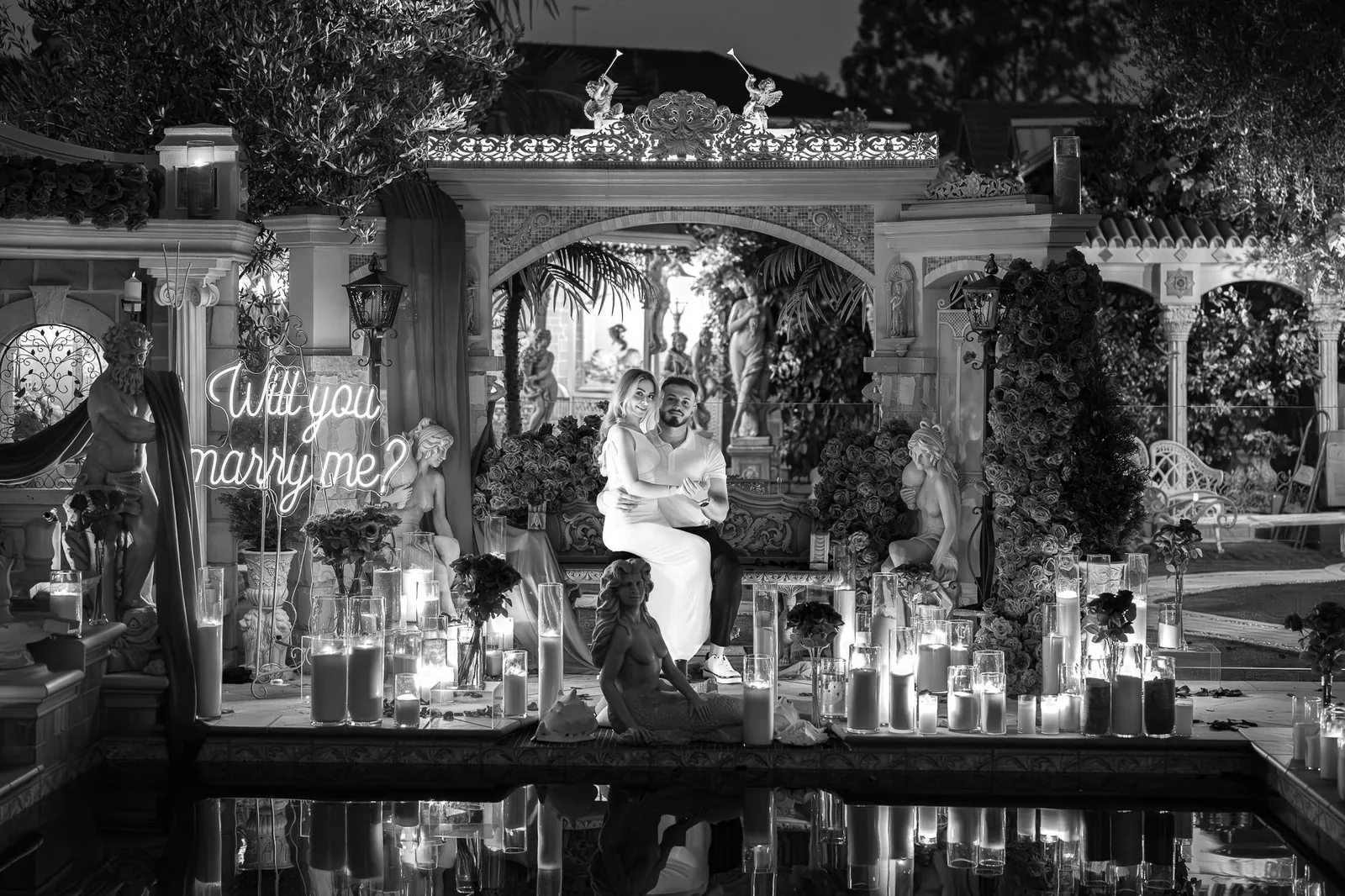 Black and white wide shot of the couple embracing in a candlelit courtyard above a reflecting pool