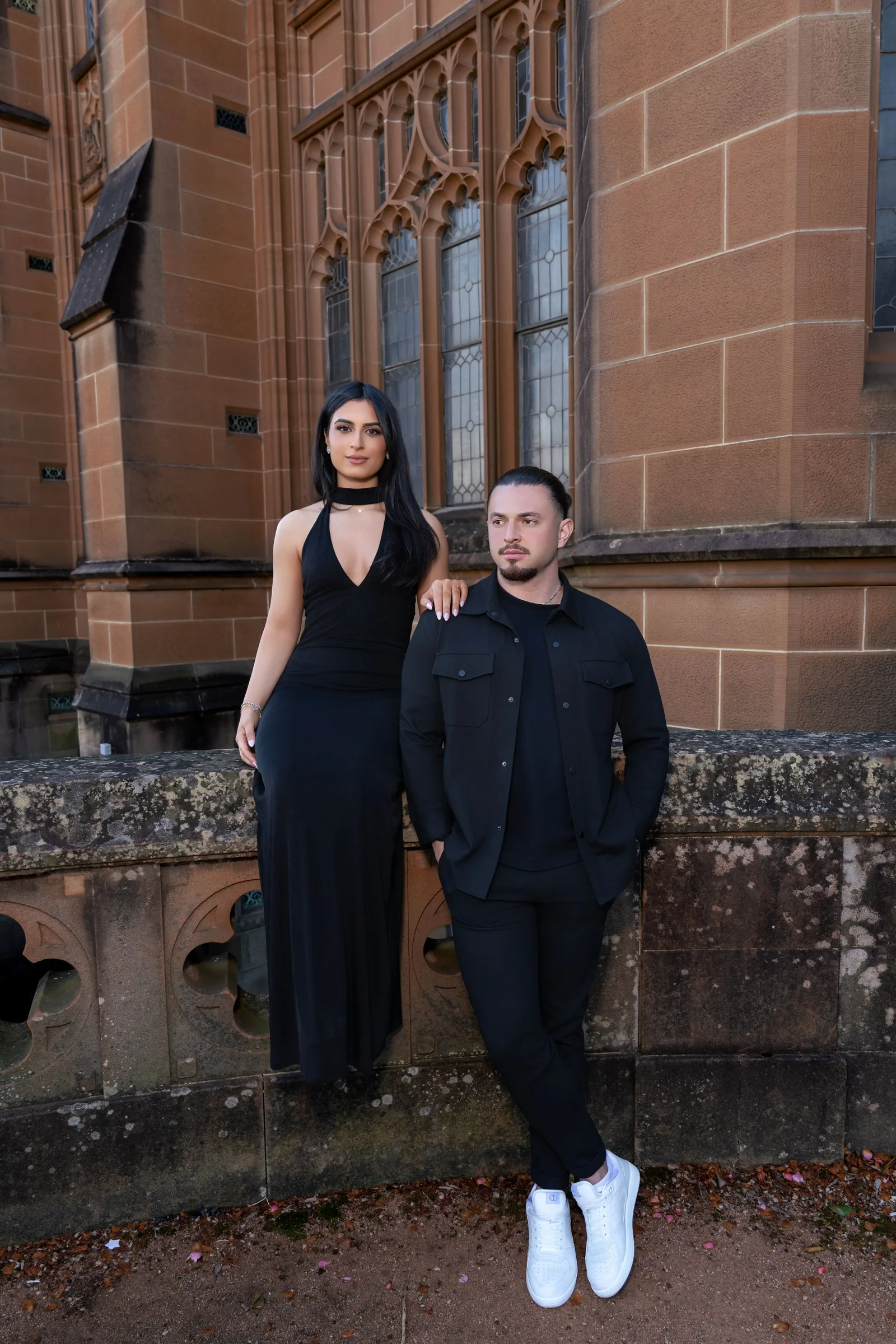 A couple in black attire poses against a sandstone church wall with gothic windows.