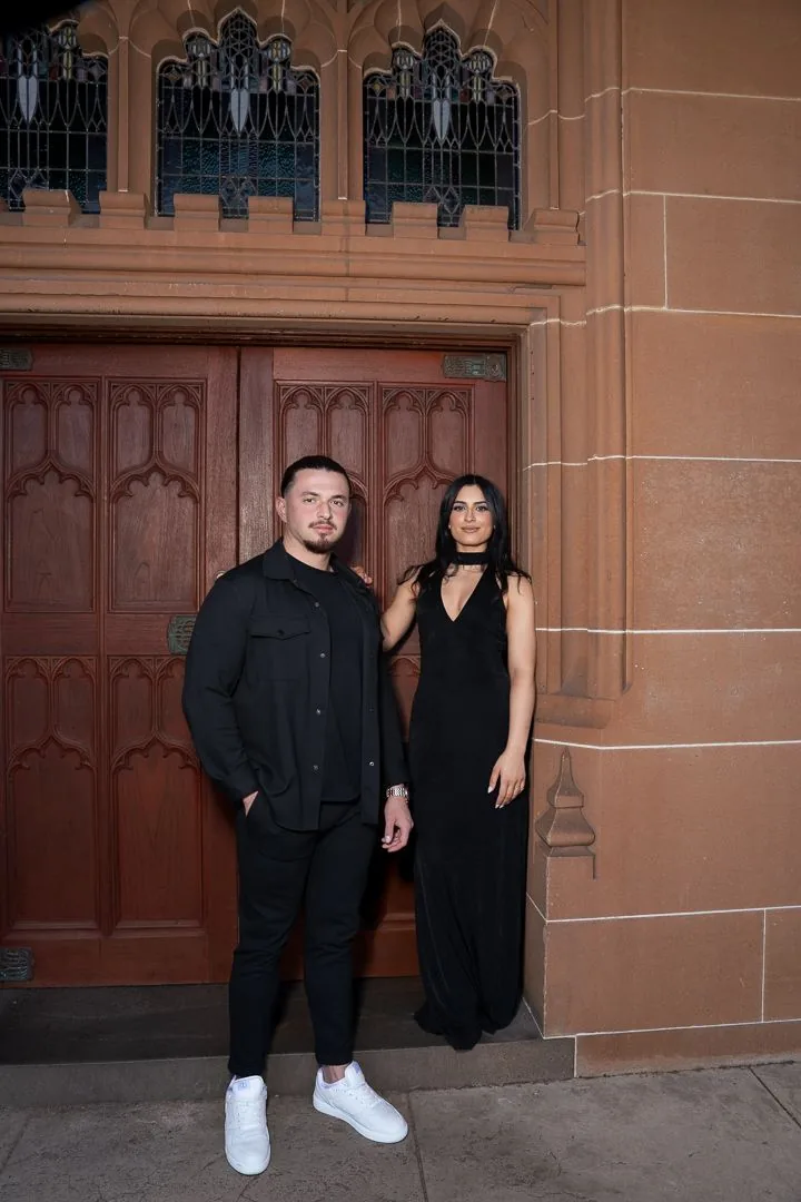 A couple dressed in black stands against ornate wooden church doors and sandstone walls.