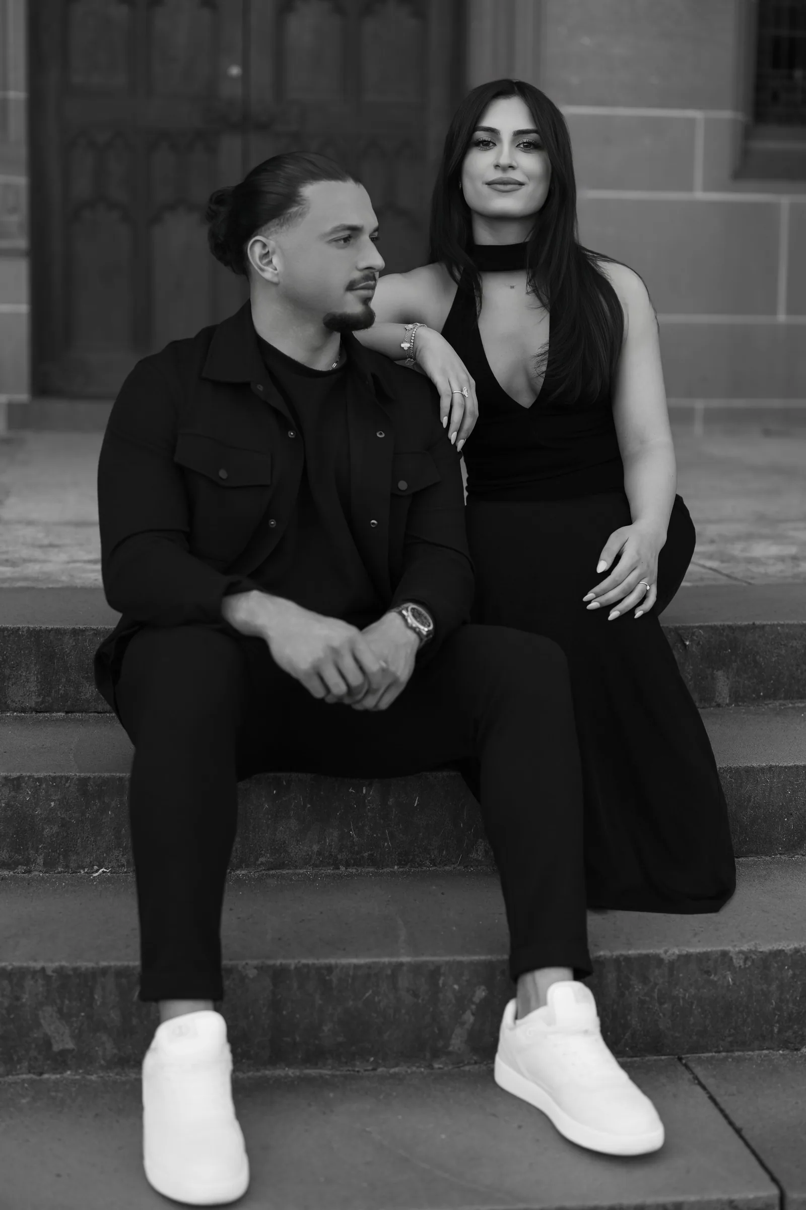 A couple in matching black outfits sits together on stone steps of a historic building.