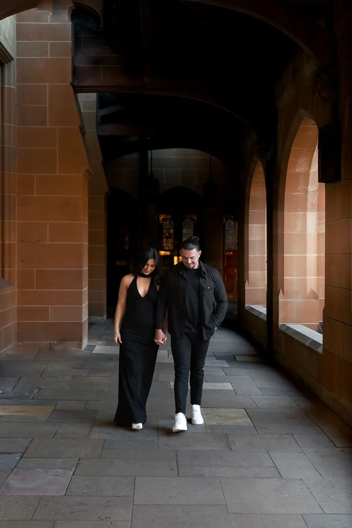 A couple walks through a dimly lit stone archway corridor in matching dark outfits.