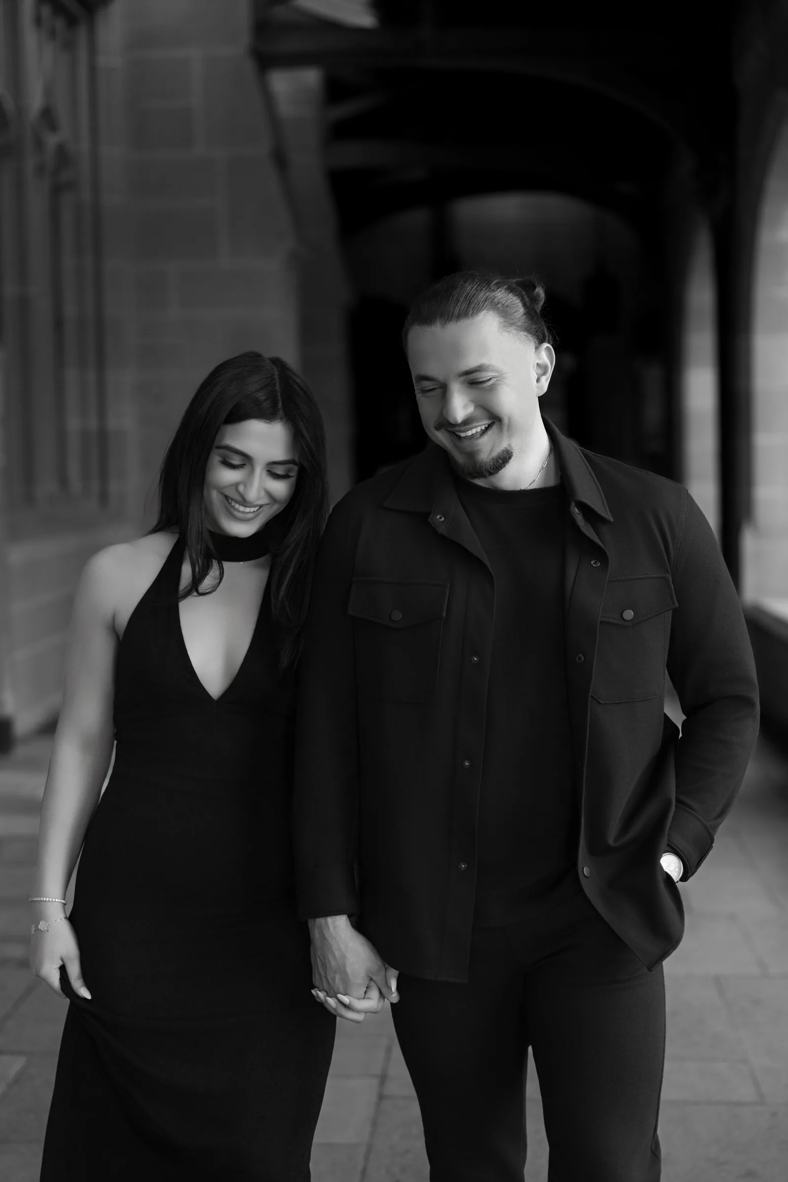 A couple in coordinated black outfits shares a tender moment while walking along a historic building.