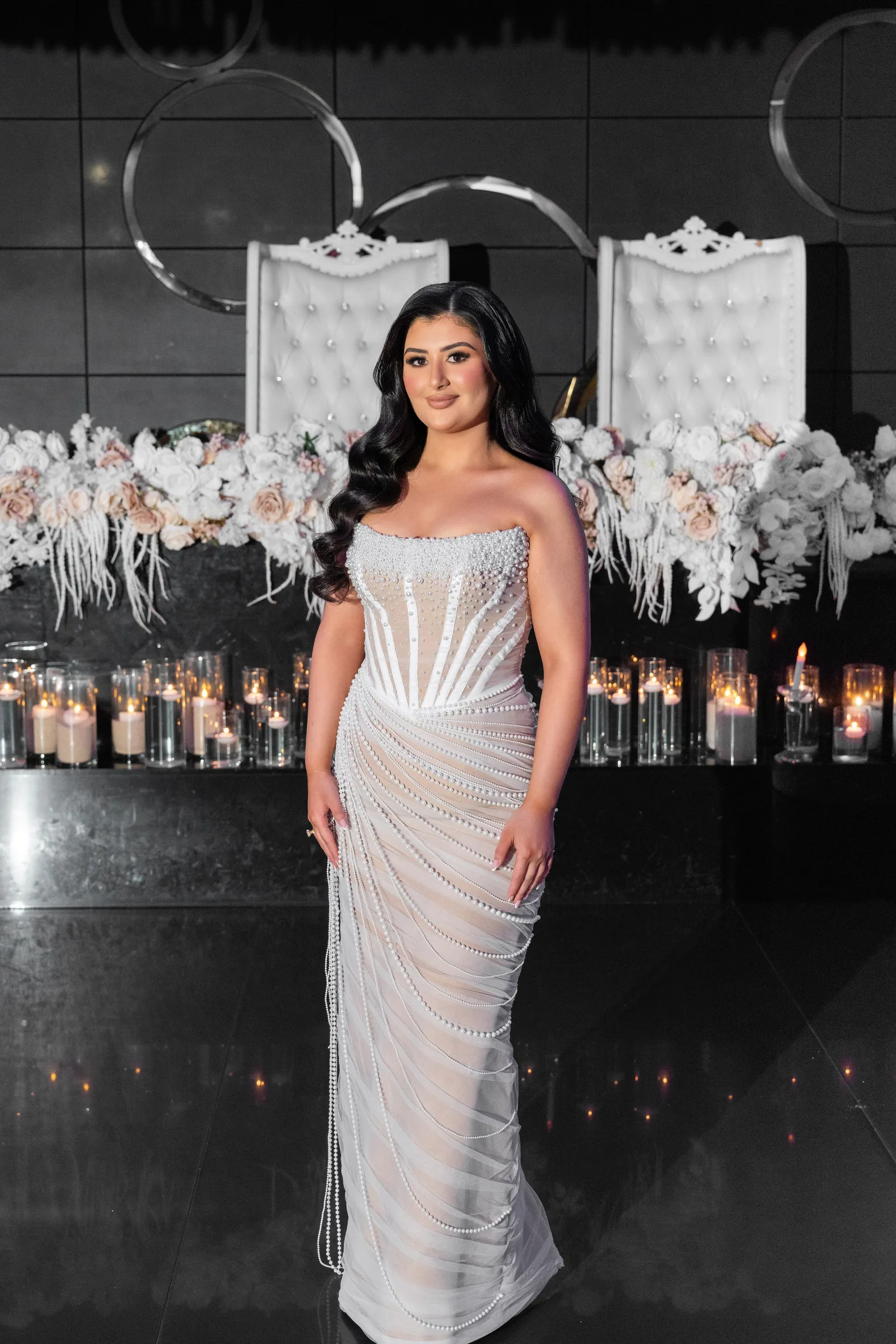 A woman poses in a glamorous white beaded gown against a dark background with hanging floral decorations.