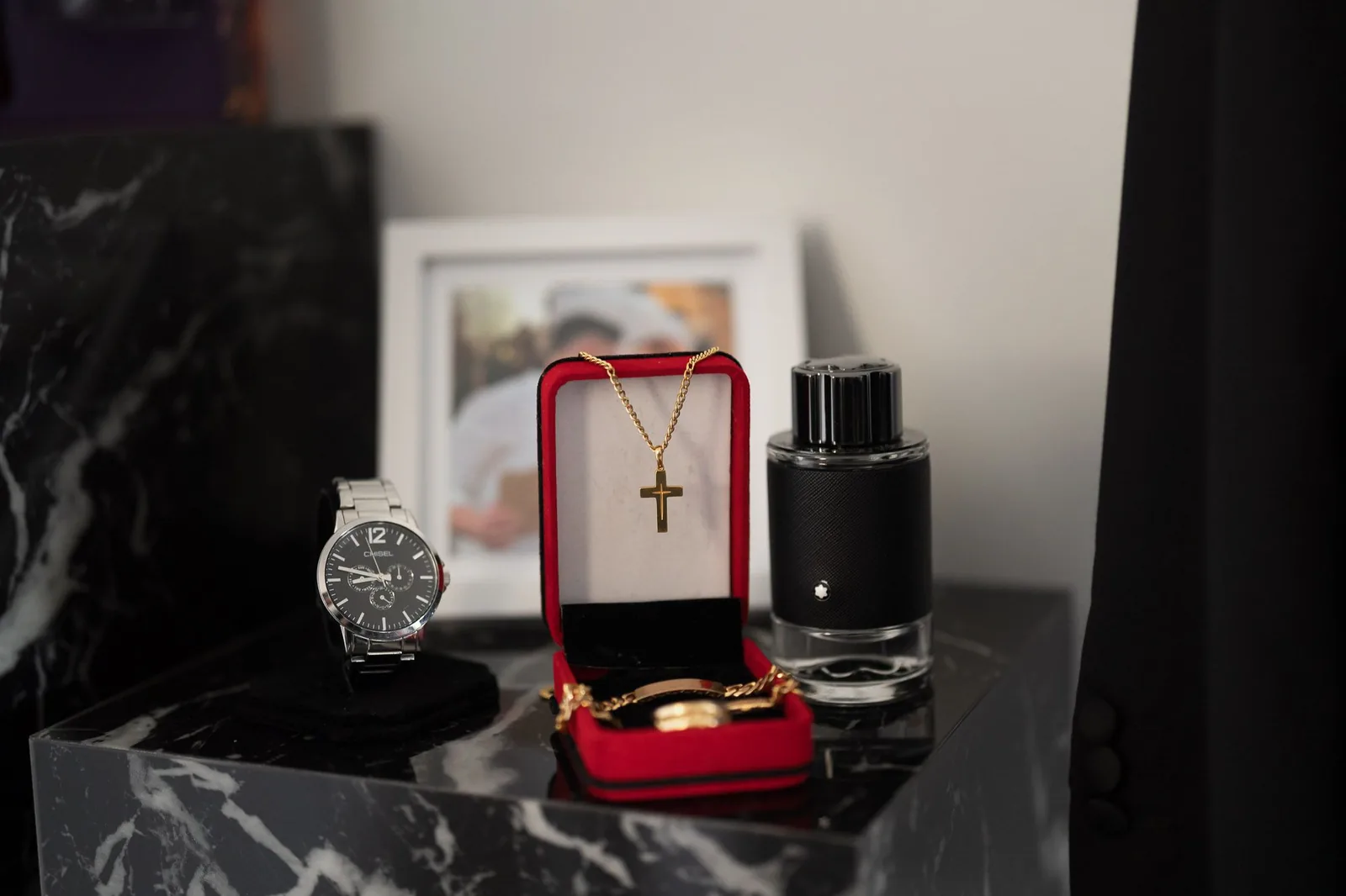 Wedding accessories including a watch, cross necklace, and rings displayed on dark marble.