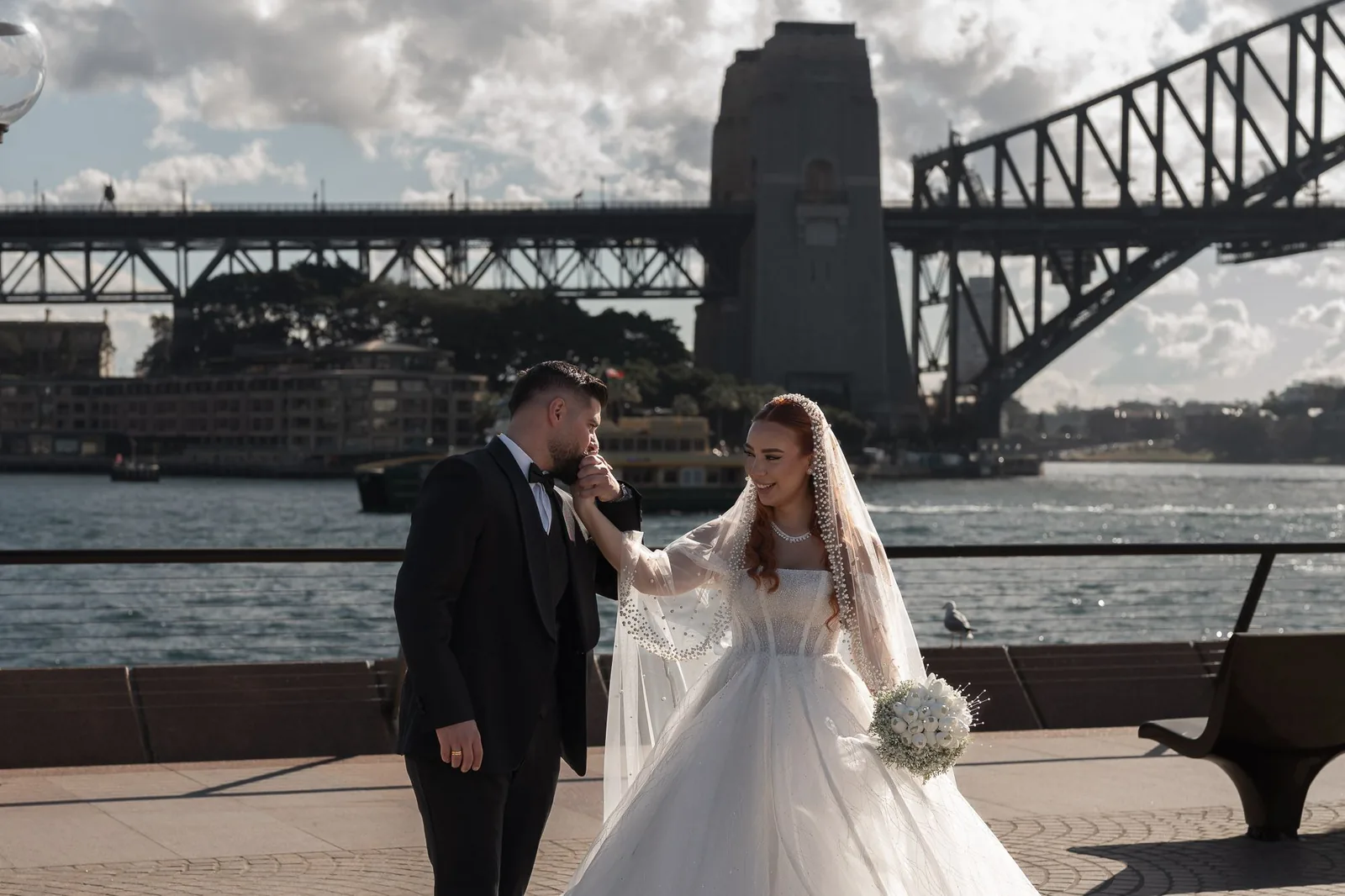 Wedding couple poses under iconic Sydney Harbour Bridge with dramatic cloudy sky.